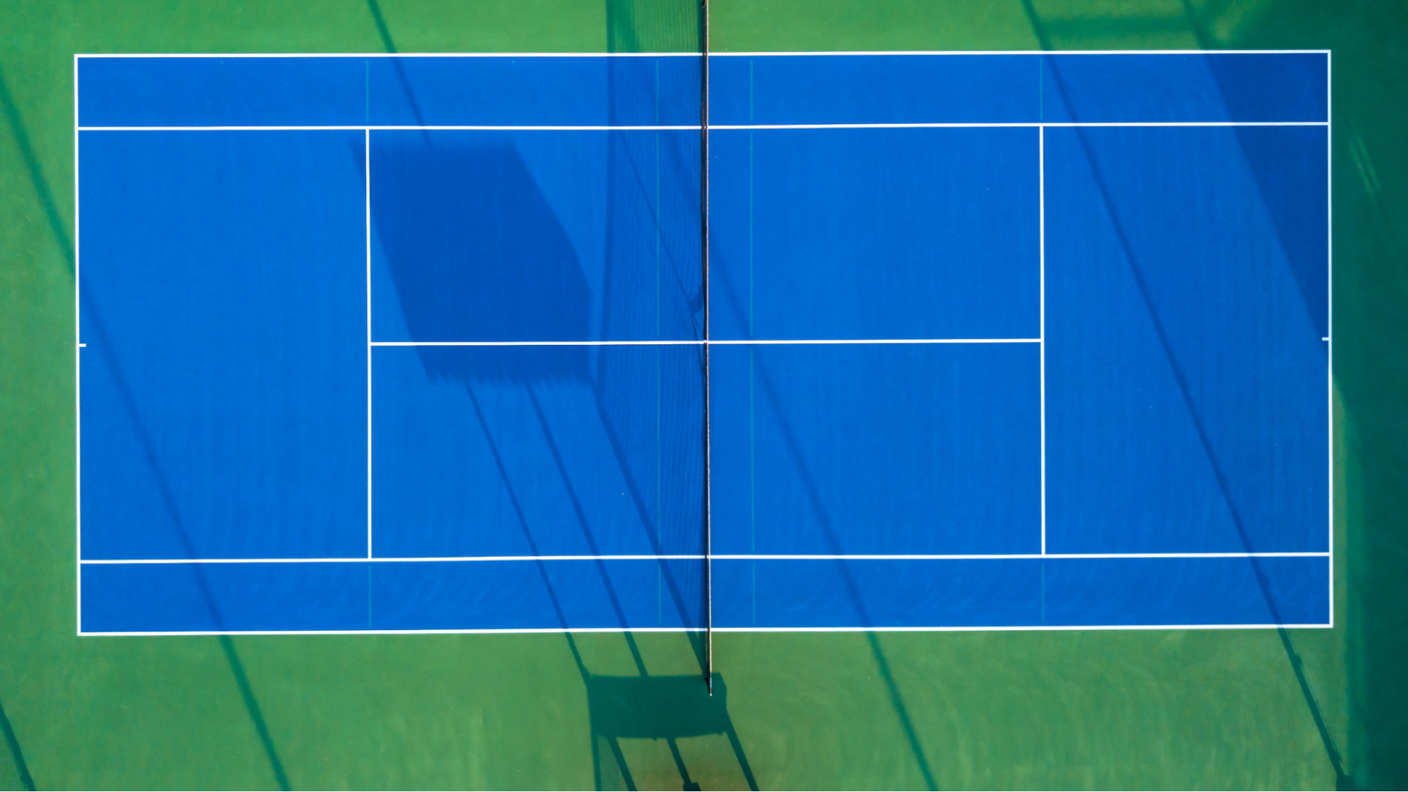 Aerial view of an empty blue Pickleball court on a green surface, casting long shadows. The image conveys a sense of symmetry and calmness.