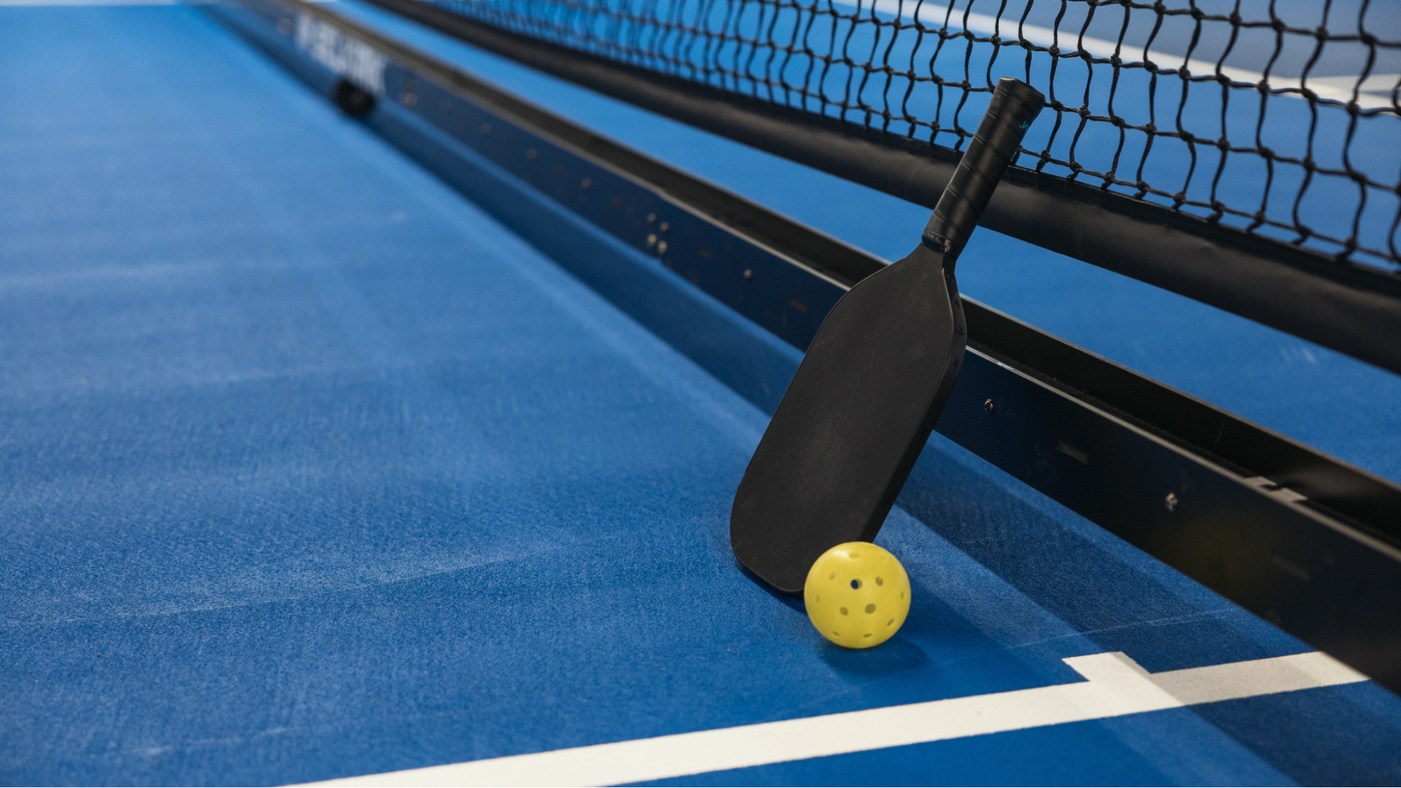A black pickleball paddle and yellow ball rest against a net on a blue court. The scene conveys a sense of calm and readiness for play.